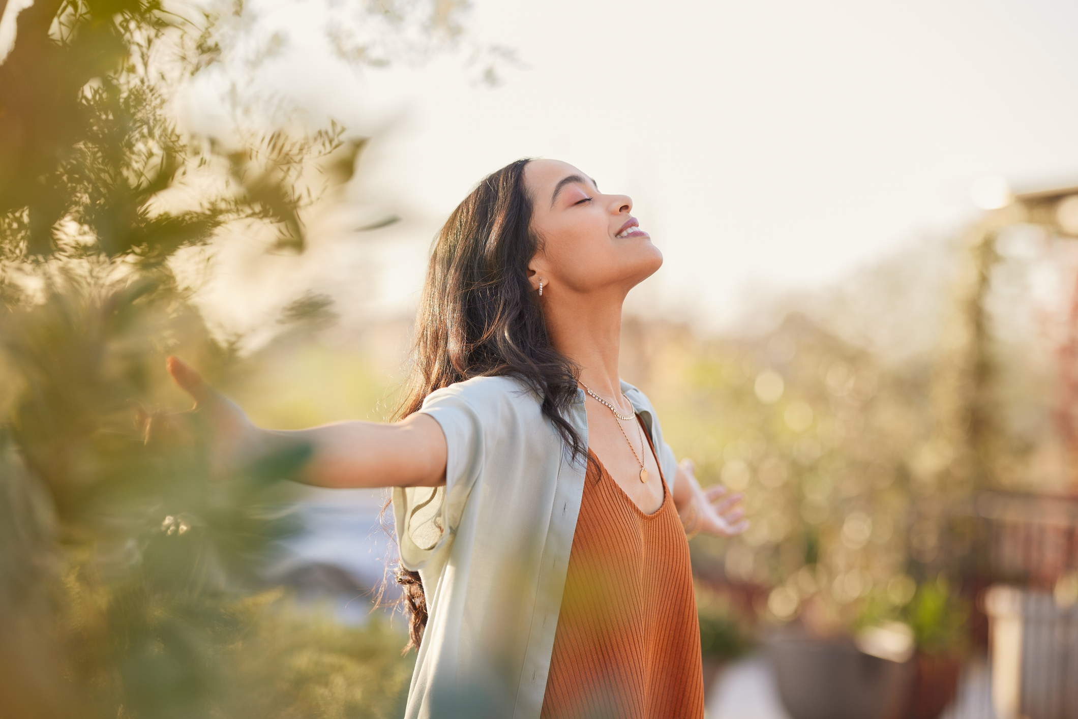 baker-12 Young latin woman with arms outstretched breathing in fresh air during sunrise at the balcony. Healthy girl enjoying nature while meditating during morning with open arms and closed eyes. Mindful woman enjoying morning ritual while relaxing in outdoor park.