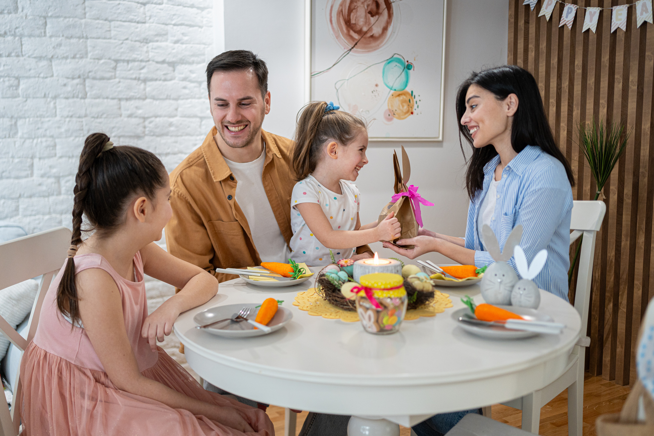 Young Caucasian family, parents with daughters celebrating an Easter, at their cozy and modern home, during an Easter lunch while exchanging Easter presents