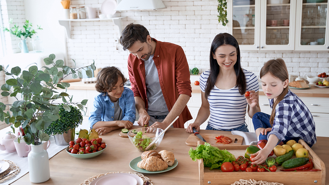 Preparing a salad. Young beautiful family cooking together in the modern kitchen at home. Mother and father teaching two little kids how to cut fresh vegetables